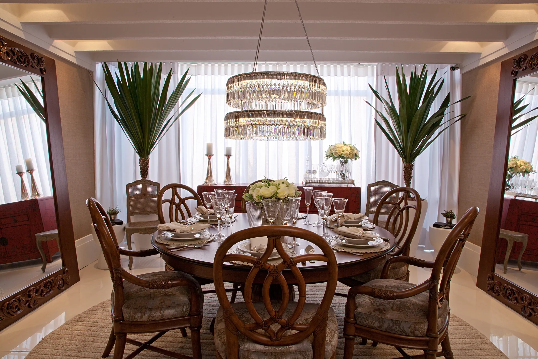 Sala de jantar com mesa redonda de madeira, cadeiras de palhinha, lustre de cristal e palmeiras em vasos