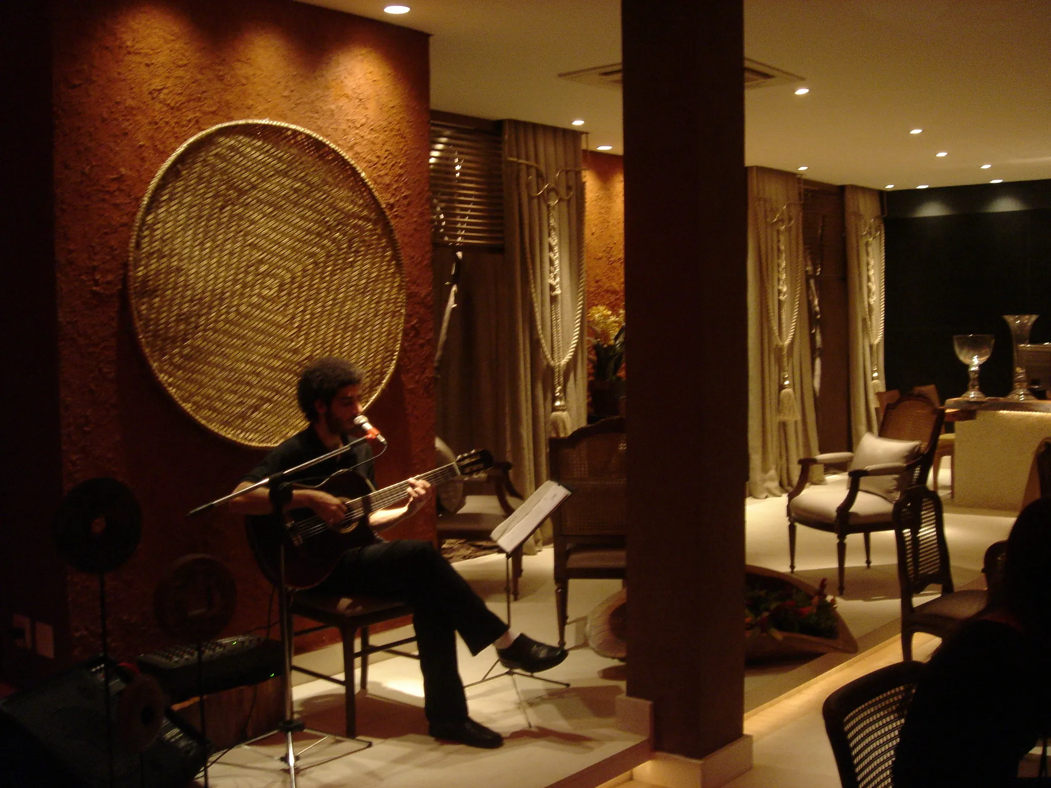 Café da mostra em linguagem colonial, com músico ao violão, peça circular de fibra na parede e mesa posta