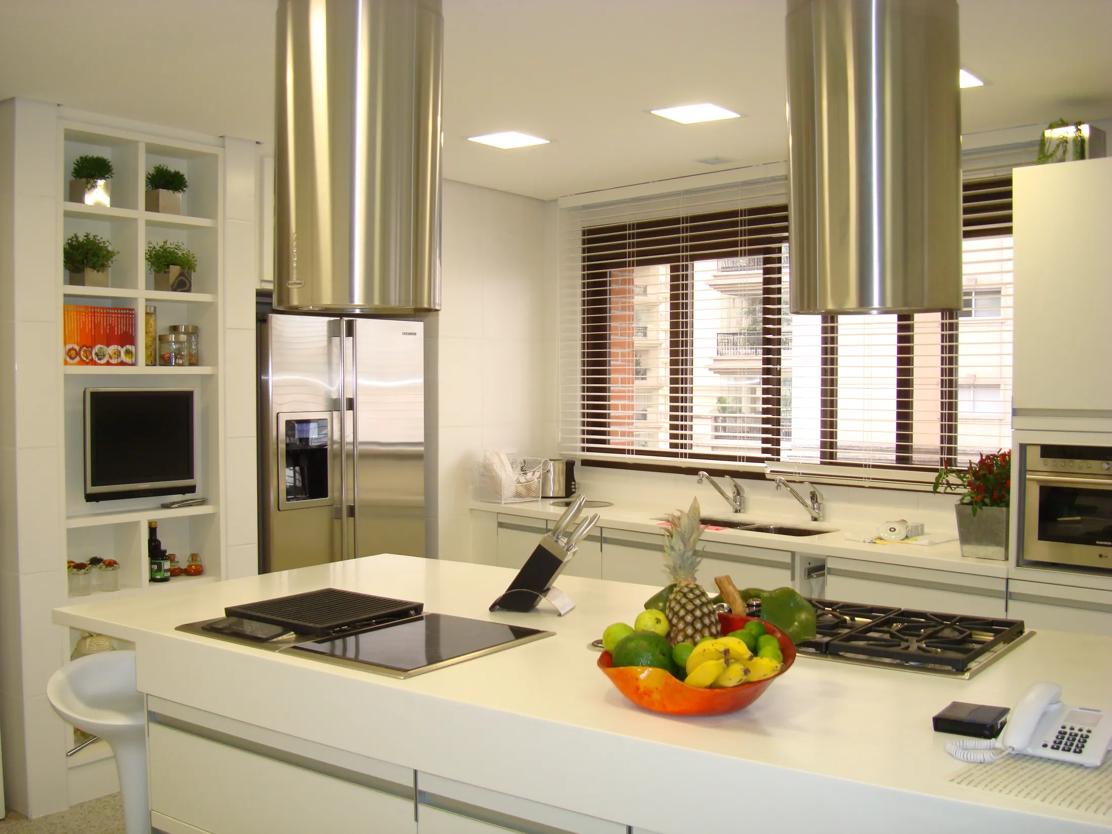Cozinha branca com ilha central, duas coifas cilíndricas em inox e marcenaria com nichos de vidro